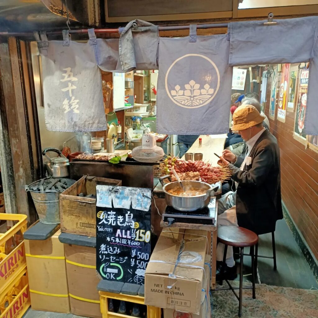 photo of a small restauarant with an older Japanese man sitting on a stool looking at his phone