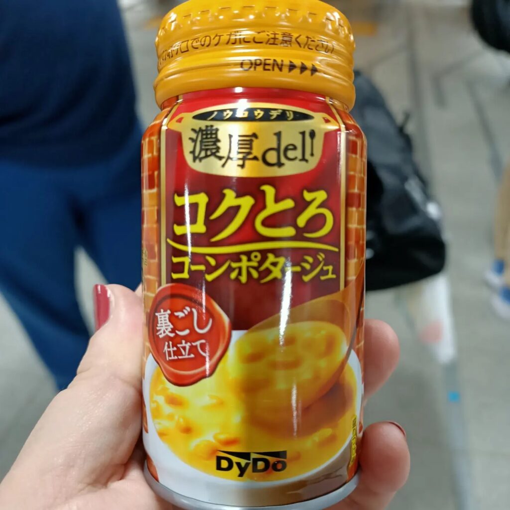 close up photo of a hand holding a small red and yellow bottle with a picture of corn soup and japanese writing