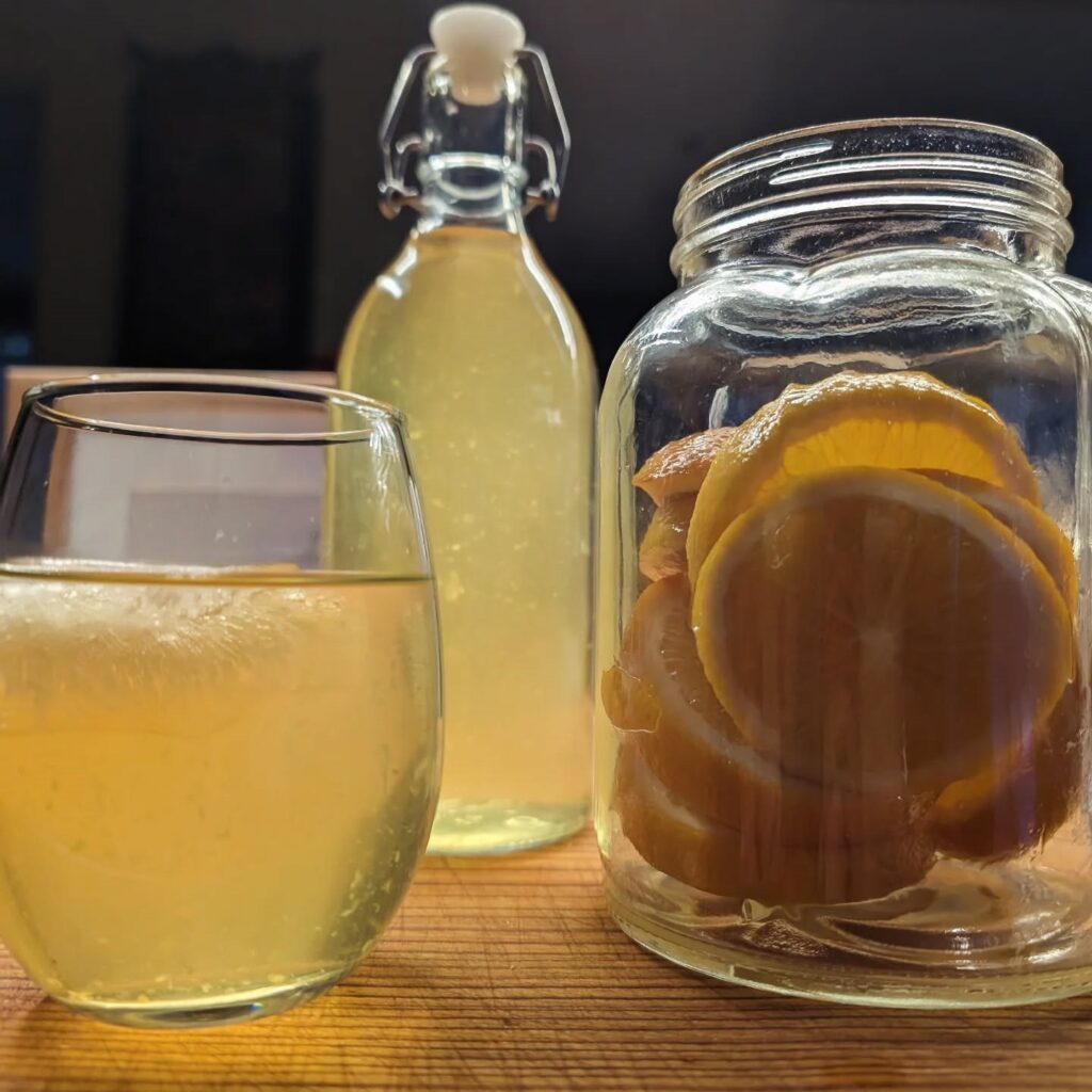 a cloudy glass of yellow orange liquid next to a bottle and a jar filled with slices of orange